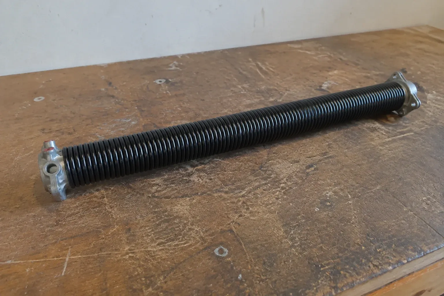 Detached black coil spring with metal end fittings on a workbench. — Torsion Spring Replacement, Haldimand County