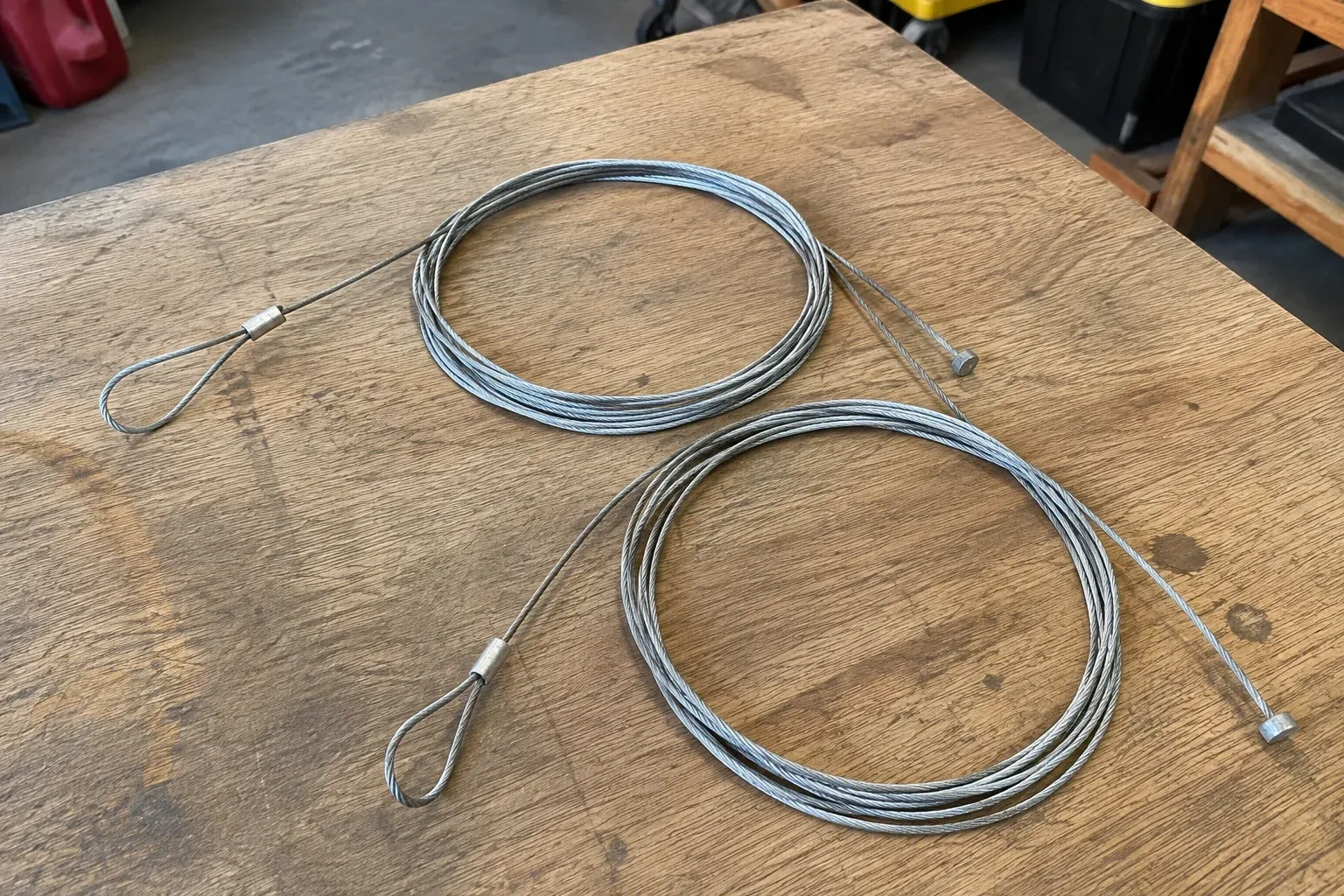Two coiled steel cables with loop and stop ends on a workbench. — Garage Door Cable Replacement, Haldimand County