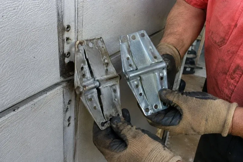 Gloved hands holding two metal garage-door hinges side by side, one discolored with a visible crack and the other bright silver. — Garage Door Hinge Replacement, Haldimand County