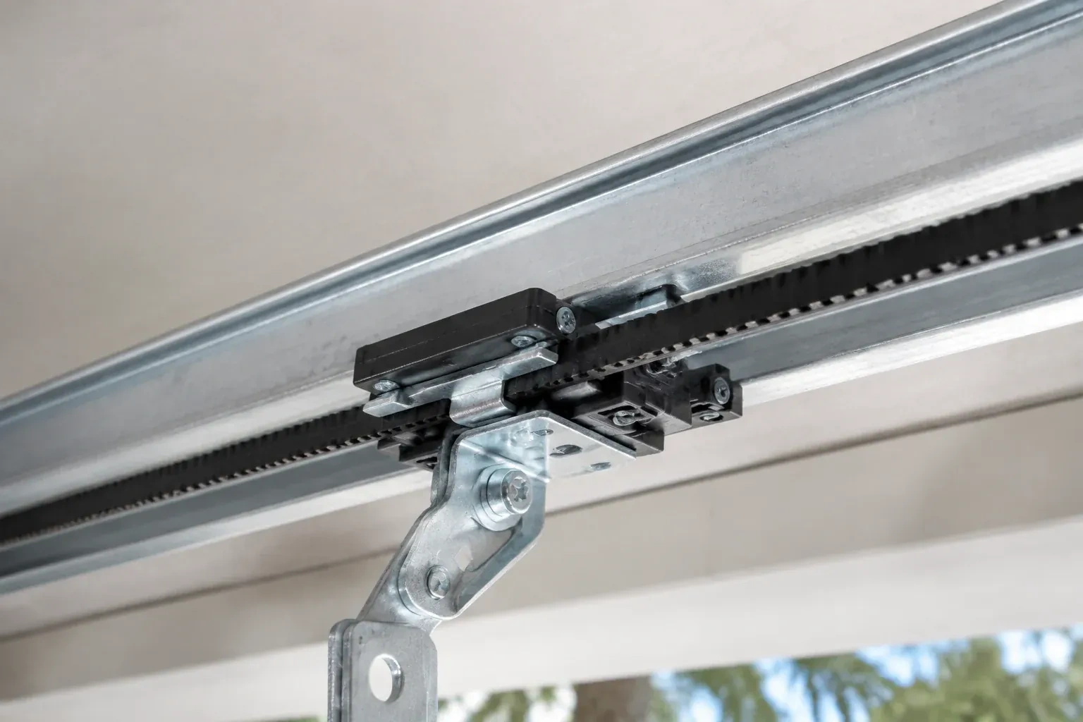 Black plastic trolley and bent galvanized bracket on a steel opener rail with a toothed black belt, against a ceiling. — Belt-Drive Opener Repair, Caledonia
